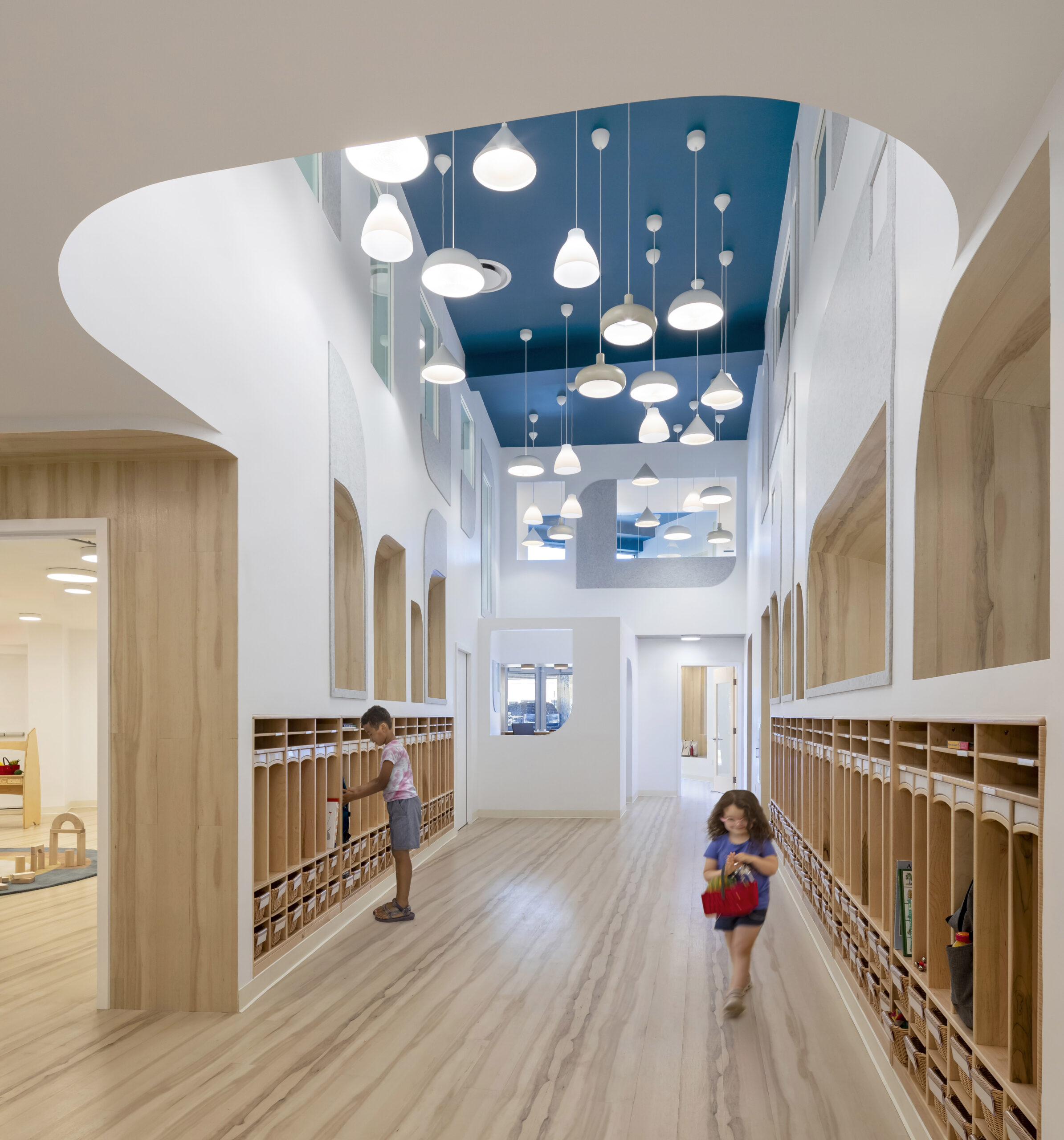 Children utilizing the convenient wooden built-in lockers in the two-story locker space, accentuated by a vibrant blue ceiling and unique lighting fixtures that create a playful and functional space.