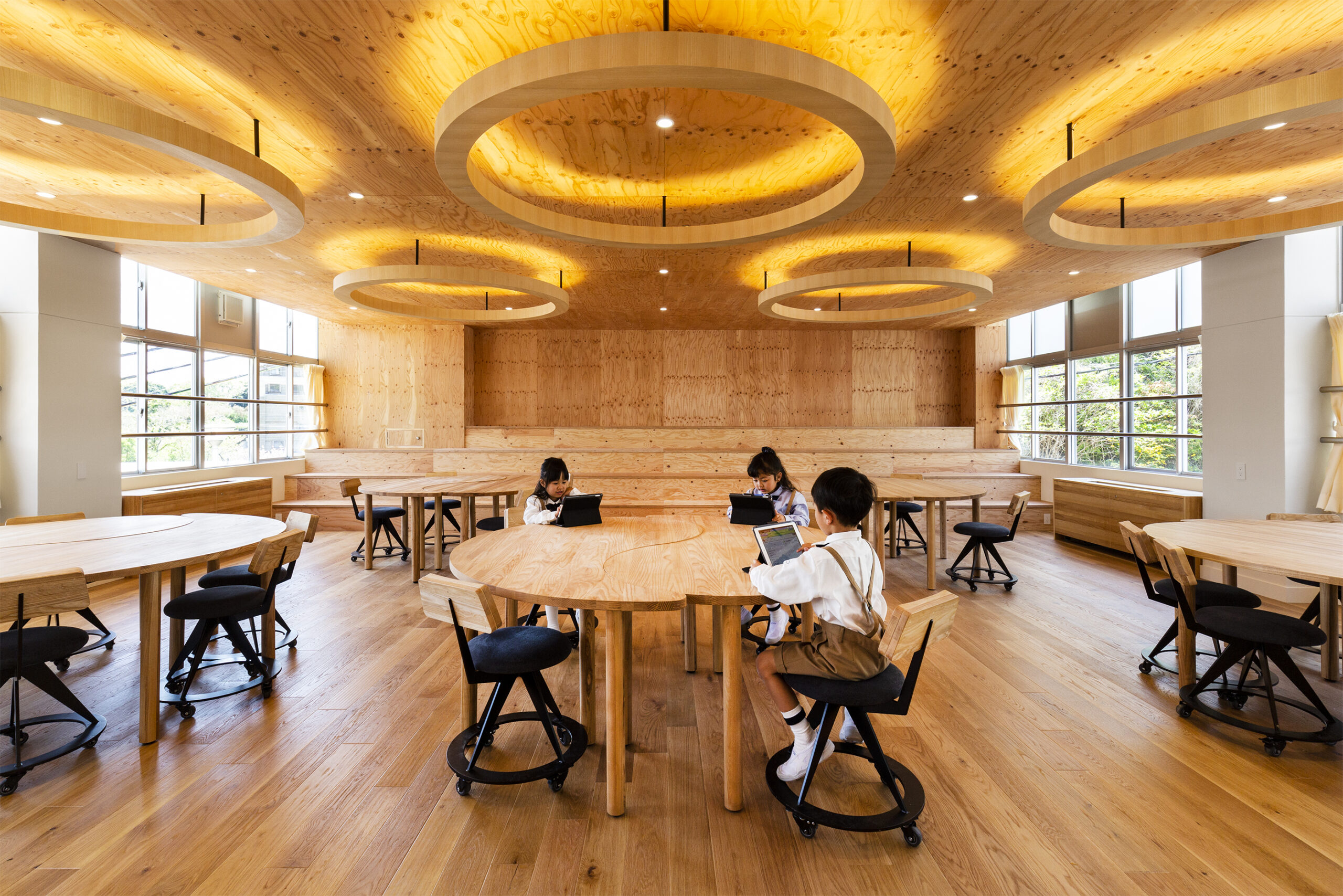 A photo showcasing a classroom, featuring wooden floors, furniture and cladding, as well as circular wooden lighting fixtures on the ceiling.